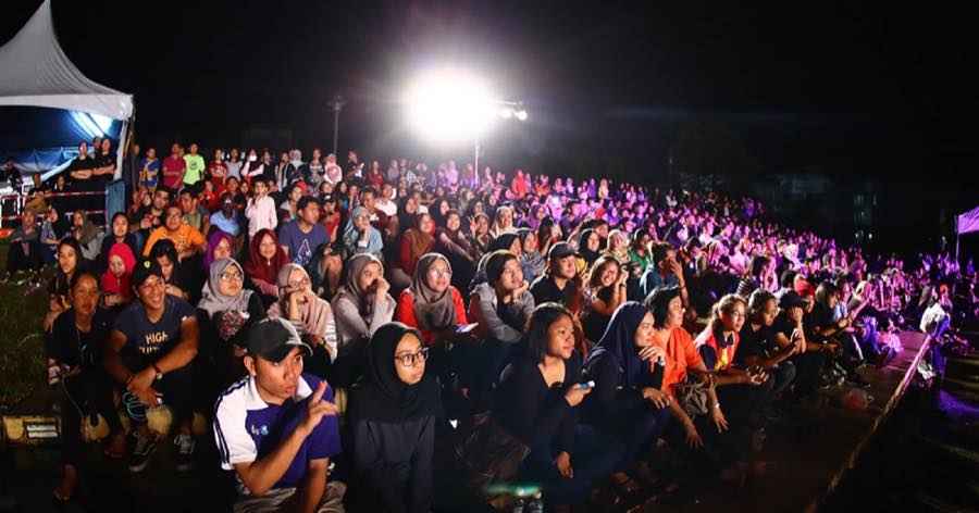 A large open-air night audience in Malaysia seated on the ground watching a lit stage