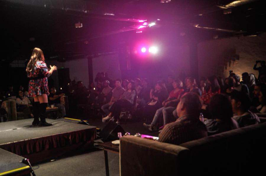A speaker on a low stage addressing a seated audience under pink stage lighting